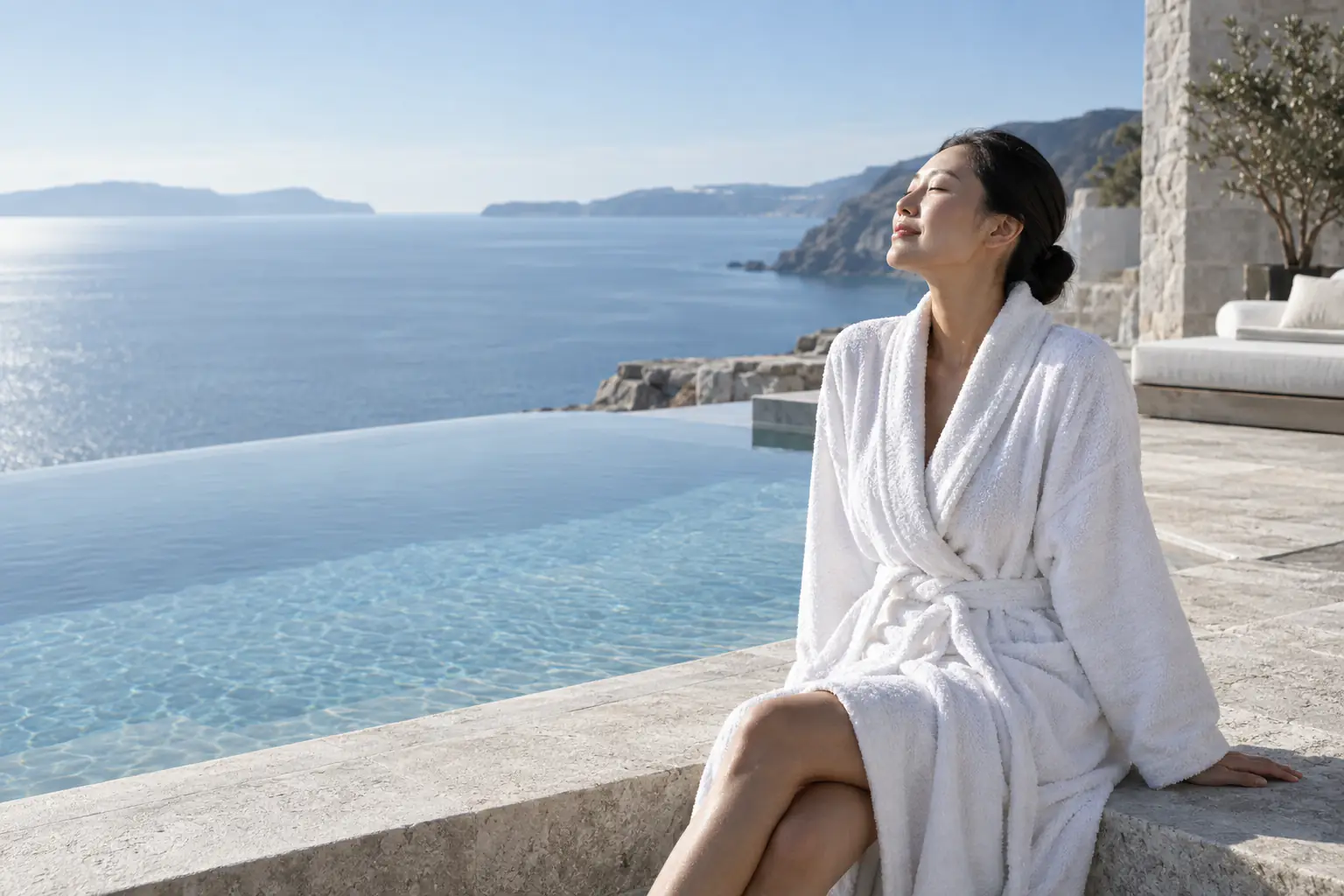 An East Asian woman in her early thirties in a white towelling robe sits at the edge of an infinity pool above the sea, face turned to the sun.