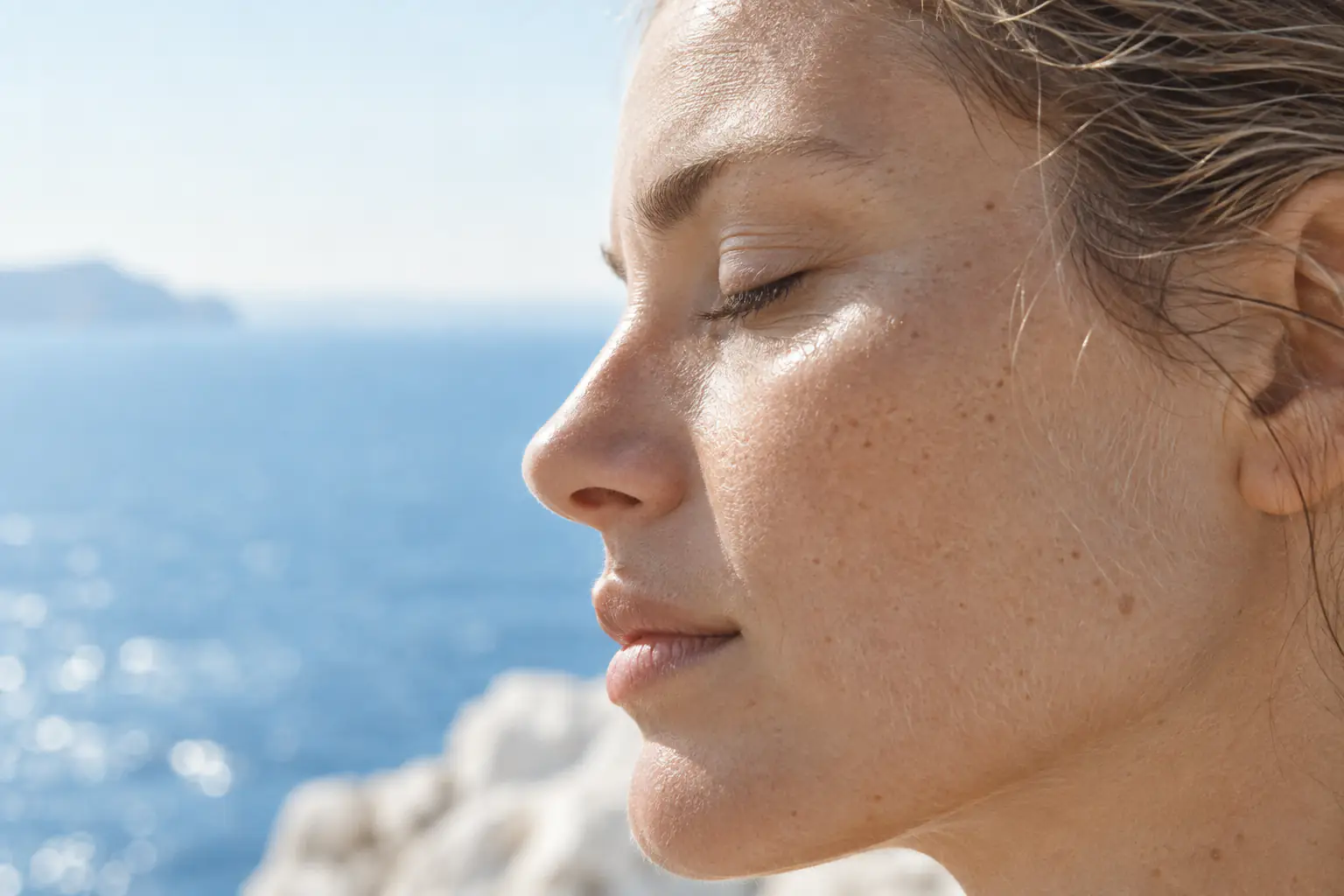 Profile of a woman in her late twenties in hard Mediterranean sun, eyes closed, the sea behind her; pores, freckles and fine facial hair in sharp detail.