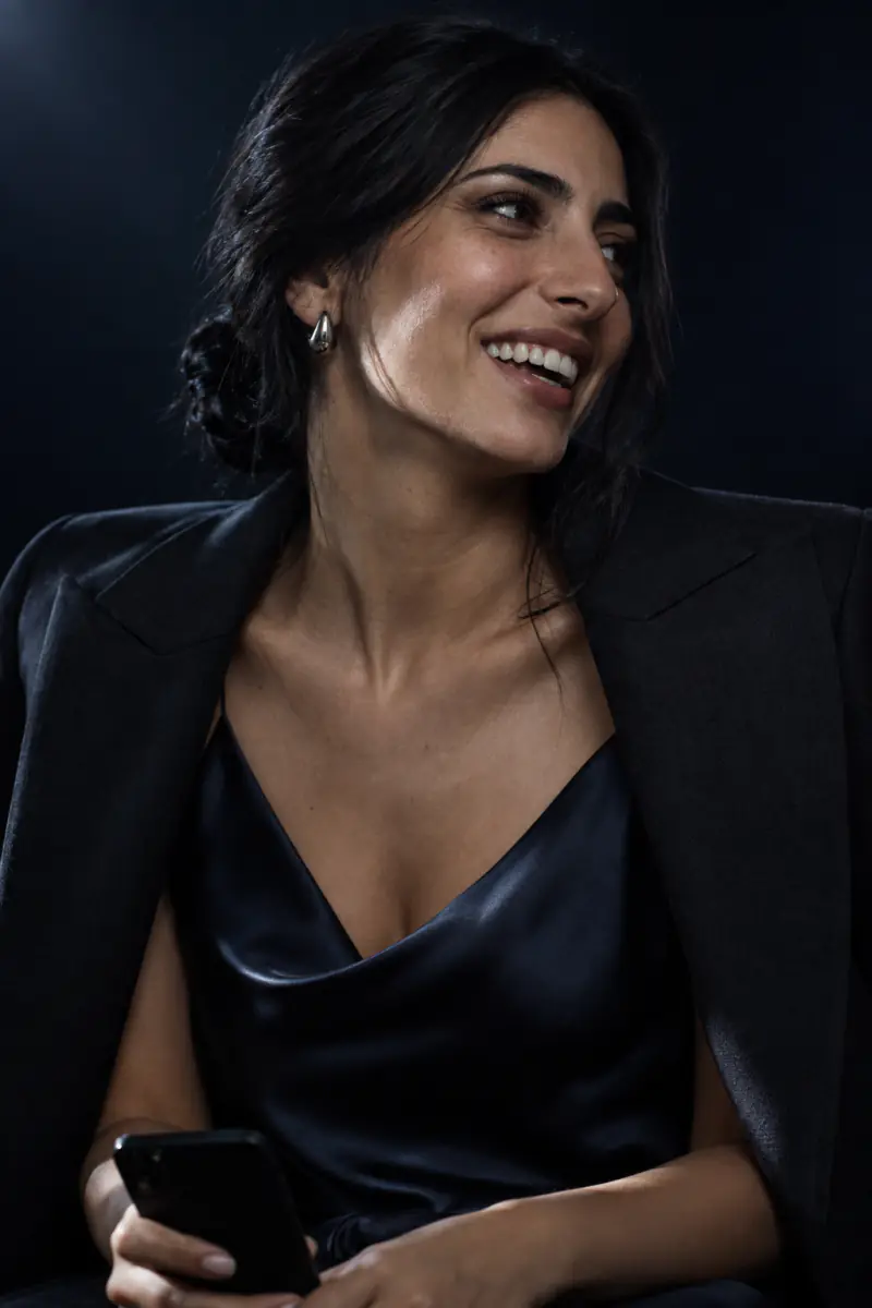 A woman in her early thirties with thick dark hair in a low knot, in a navy satin slip and charcoal jacket, laughing with her phone in her hand.
